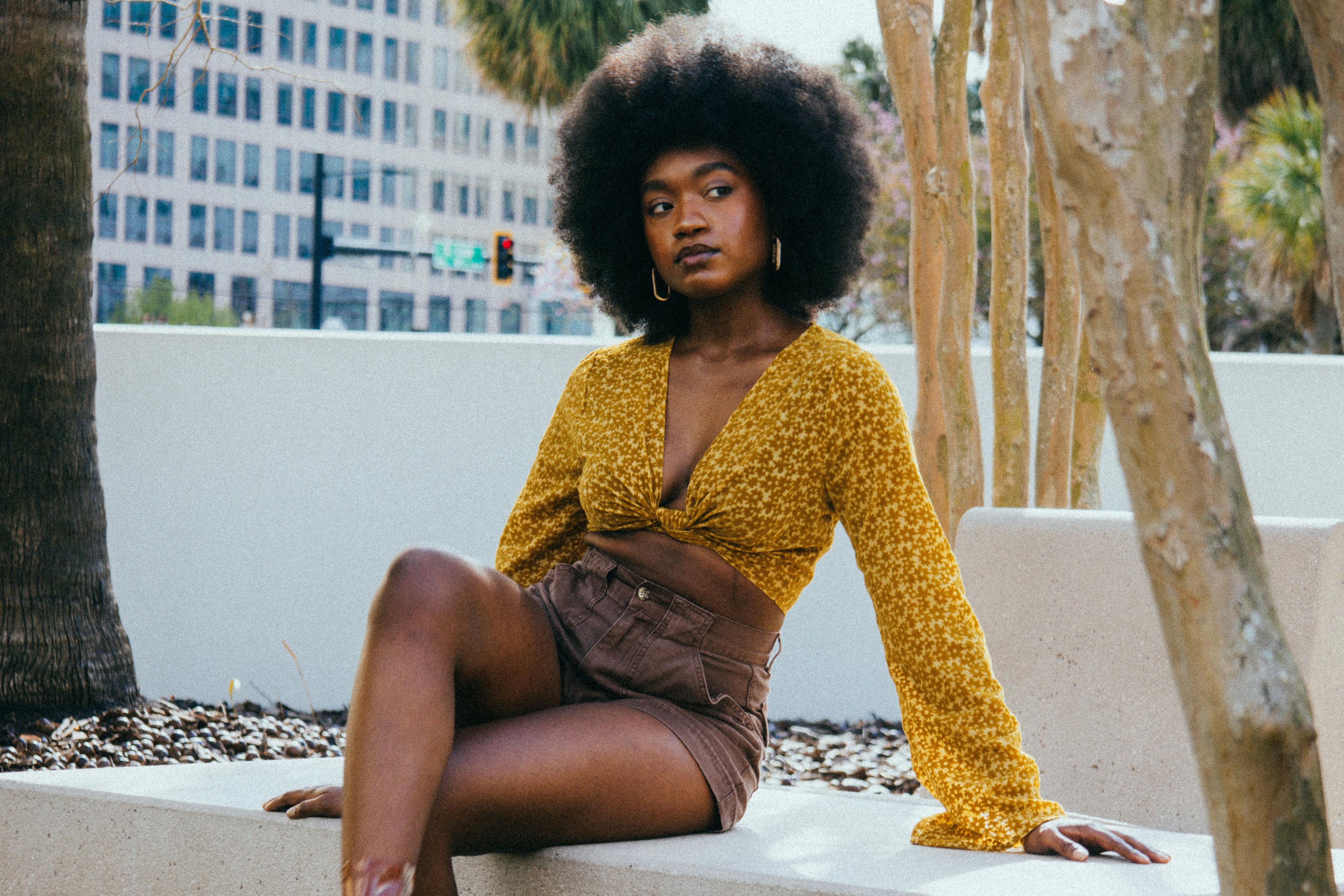 A young lady sits on a concrete bench. She is sitting with her legs crossed and leaning back on her hands. Her eyes and body language are shifted to the right.