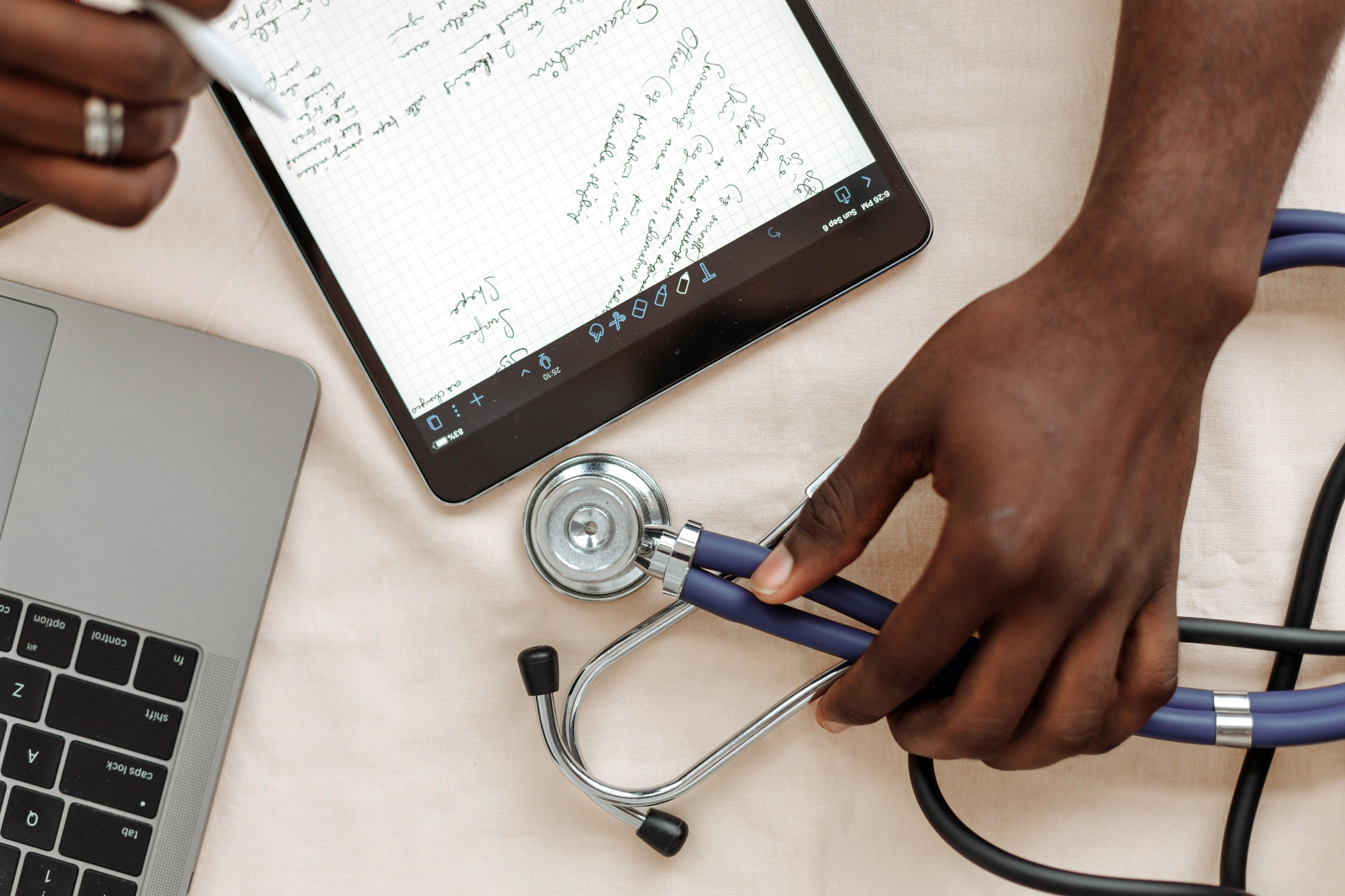 A doctor's hand rests on a stethoscope sitting on a table. On the table, there is also a laptop and an ipad with notes.